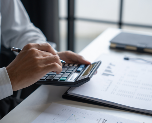 Close-up of a person's hands using a calculator and pen on a desk with financial documents and graphs.
