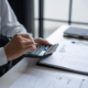 Close-up of a person's hands using a calculator and pen on a desk with financial documents and graphs.